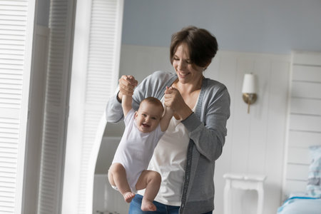 Cheerful young mom playing with cute baby, holding kids hands, lifting hanging infant in air, playing with little child, training muscles, laughing, having fun, enjoying maternity leaveの写真素材