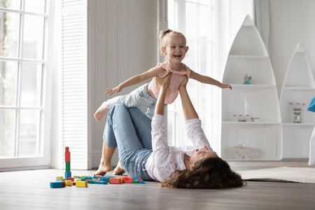 Cheerful excited kid girl and happy mom playing airplane flight, doing acroyoga support, having fun on heating floor. Mum lying on back, holding daughter, lifting child up in air, laughingの写真素材