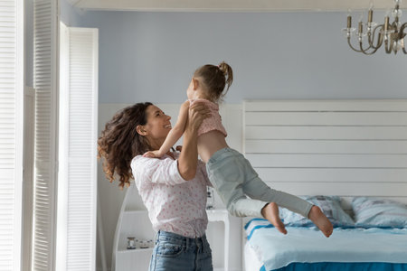 Happy energetic mother playing active games with little kid at home, lifting girl up in air, smiling, laughing, enjoying motherhood, family activity, leisure, having fun, dancingの写真素材