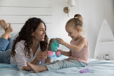 Cheerful playful mom and cute little kid playing role game, tea party, coffee shop on bed, pretending pouring drink into toy dish, smiling, laughing, having fun, spending leisure time, weekend at homeの写真素材