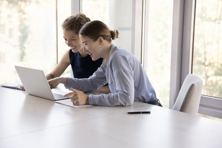 Two cheerful young female employees sharing workplace, using online application on laptop, watching presentation on Internet together, smiling, laughingの写真素材