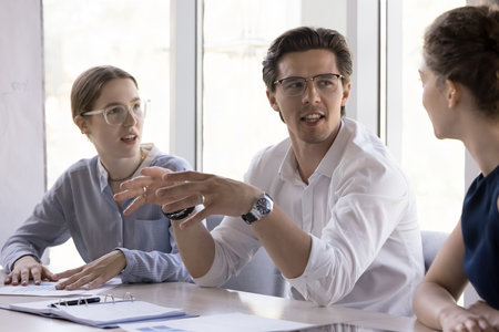 Confident handsome young professional man speaking to female colleagues on brainstorming meeting,...