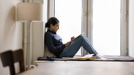Indian woman spend leisure use digital tablet, rest on windowsill at home, websurfing information, make order, do e-shopping through retail on-line services. Modern tech for study, leisure, apps usageの写真素材
