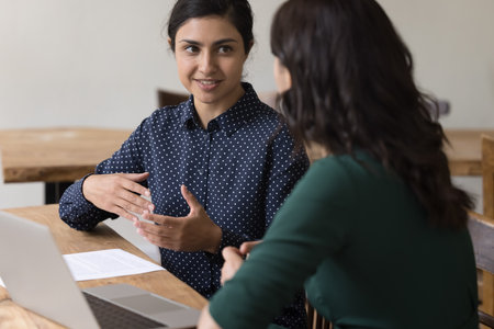 Two young businesswomen, colleagues or students discuss joint task seated at desk with laptop....
