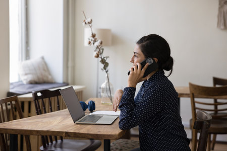 Indian businesslady talks on smartphone sit at desk with laptop, lead formal conversation, ask or provide information, communicates to customers support services use modern tech and mobile connectionの写真素材