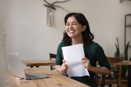 Happy young woman sit at home workplace hold paper letter, received bank loan approval notice, job promotion or salary increase, surprised by great news, money refund document, enjoy moment of successの写真素材