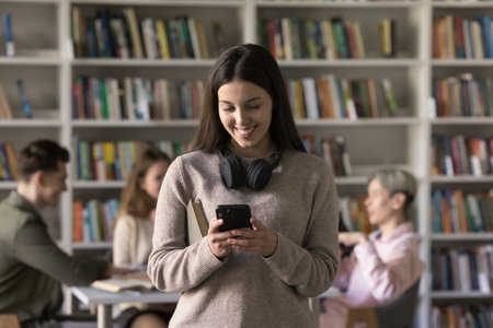 Teenage student girl standing in library or classroom use smart phone, search information, take break, read sms from friend, chatting on-line, check timetable through mobile apps. Modern tech, studiesの写真素材