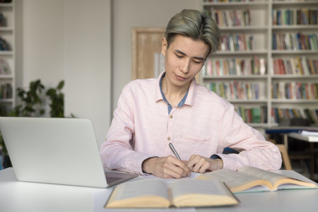 Serious Asian student guy sit at desk in library, use laptop, work on project, write notes, do homework, prepare for session, use internet resource website, get ready for admission, gain new knowledgeの写真素材