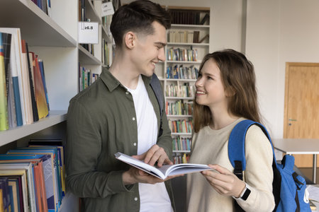 Two smiling pretty girl and guy with textbook standing in library, prepare for exams together,...