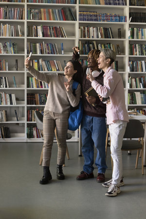 Three multi ethnic friends, girl and guys students standing in library staring at cellphone screen make selfie pictures, gesturing, record video for channel. Generation Z and modern tech usage, funの写真素材