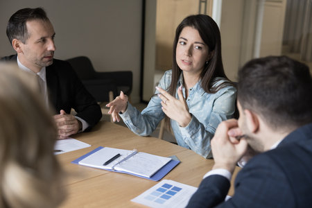 Confident business leader woman talking to colleagues on corporate meeting, speaking at conference table, discussing teamwork. Young businesswoman brainstorming on project with coworkersの写真素材