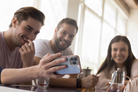 Guy holds cellphone showing to friends new mobile application, game, watch video spend time together on internet gather in cafe drink tea enjoy funny amusing on-line content. Fun, leisure, modern techの写真素材