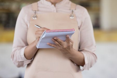 Crop midsection close up view, unknown waitress in apron hold pen and notepad ready to take order standing at workplace. Good customer services, food and drink commercial establishment worker workflowの写真素材