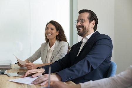 Millennial Caucasian and Hispanic businesspeople listening presentation, laughing seated at desk in...