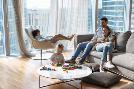 Couple of happy young parents resting on sofa in city apartment with large window, watching children, Two kids enjoying leisure at home, constructing toy building blocks, relaxing in armchairの写真素材