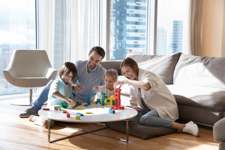 Happy positive parents and two little sibling children building towers from colorful blocks, playing game for architecture skills training, development, sitting on floor in living roomの写真素材