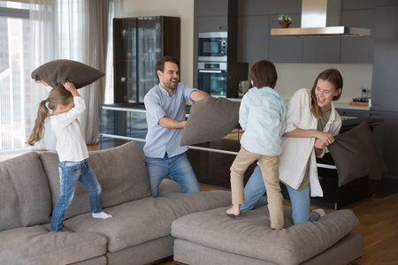 Energetic little kids and cheerful parents playing active games at home, waving pillows, fighting on big soft couch, having fun, laughing, enjoying family leisure, playtime, parenthood, childhoodの写真素材