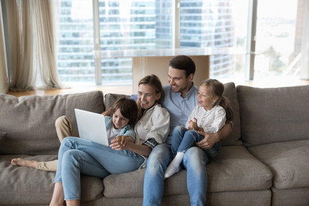 Happy little children and cheerful mother and father watching funny family movie, using learning app on laptop, enjoying leisure time on home couch, using computer, digital gadget for communicationの写真素材