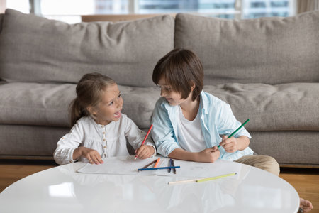 Cheerful little sibling children enjoying creative hobby, leisure, daycare activity at home together, chatting, talking, drawing with pencils, discussing colorful graphic doodlesの写真素材