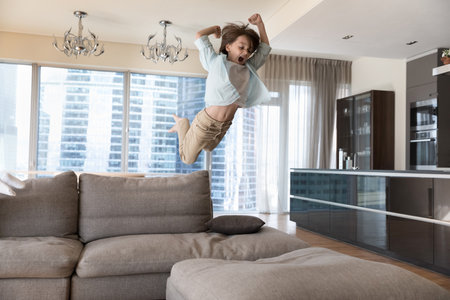 Excited crazy boy kid jumping on big sofa, throwing body on soft couch with wild scream, flying in air, playing game, enjoying activities, leisure time, playtime at home, having funの写真素材