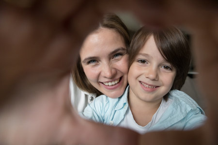 Happy positive pretty little son and young mother looking at camera through hand heart finger frame, smiling, laughing, having fun, showing symbol of love, posing for family closeup portraitの写真素材