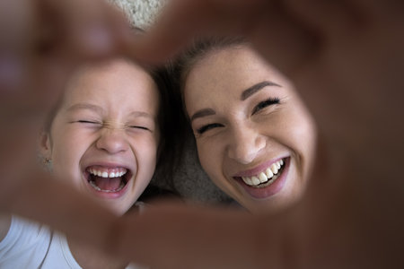Happy excited Asian little kid girl and young mom showing hand heart, looking at camera through finger frame, smiling, laughing, having fun, lying on floor, posing for close up shotの写真素材