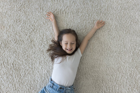 Cheerful cute little Asian kid resting on heating clean carpeted floor, lying on back with closed open arms, smiling, laughing, enjoying leisure, playing in cizy safe home. Top view portraitの写真素材