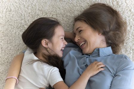 Happy positive young Asian mother and cute little kid girl lying on warm soft floor, hugging, chatting, talking, smiling, laughing. Loving caring mom caressing sweet preschool childの写真素材