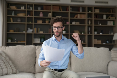 Happy excited young man in glasses reading paper letter at home, receiving document, making victory...
