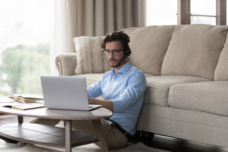 Focused positive young adult student man in wireless headset using laptop, open books for online education from home, speaking at learning conference, watching webinar, talking on video callの写真素材