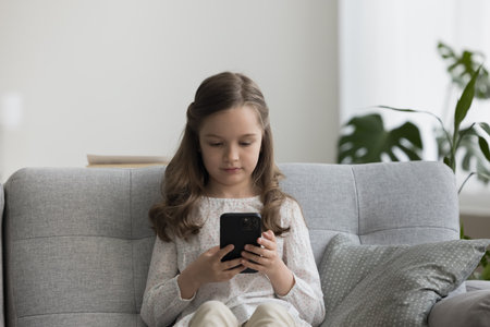 Cute girl sit on couch in living room looks at smartphone screen, play on-line game, watch cartoons, makes video call, chatting with friend, communicate to parents through messenger, using mobile appの写真素材