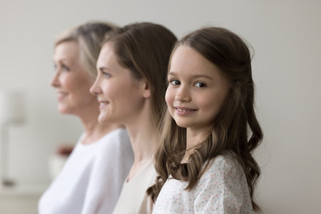Happy preschooler girl staring at camera posing standing in row with young pretty mother and older...