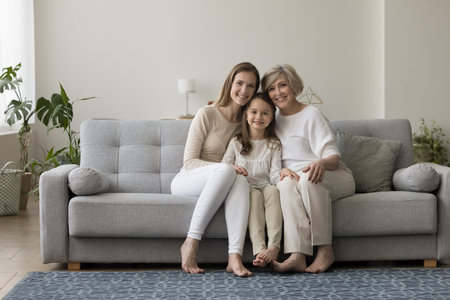 Three generations of women sit on sofa smile look at camera. Elderly 60s grandma her cute little grandchild and grownup daughter shooting for picture in living room. Multigenerational family portraitの写真素材