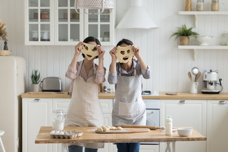Cheerful playful young adult daughter and mature mom in baker aprons having fun, laughing in home kitchen, holding funny face masks from raw dough, posing for camera, cooking, bakingの写真素材