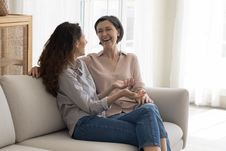 Cheerful senior mature pretty mom and adult daughter woman meeting at home, talking, discussing good news, family gossip, sitting close, speaking, laughing with joyの写真素材