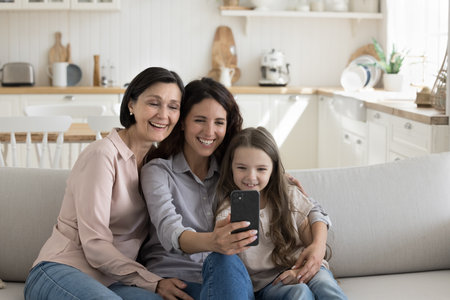 Cheerful girls and women of three family female generations using mobile phone, taking selfie on...