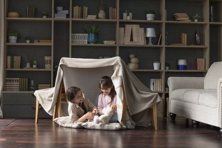 Happy mom and adorable little child girl having fun in play tent, resting on heating floor in handmade toy fort from chairs and blanket, talking, chatting, laughingの写真素材