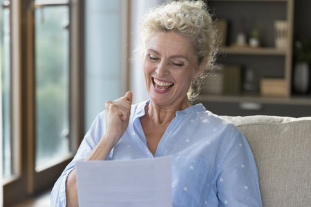 Cheerful happy mature woman reading paper letter, holding legal document, contract, retirement insurance agreement, making winner hand fist, winning prize, laughing, smilingの写真素材