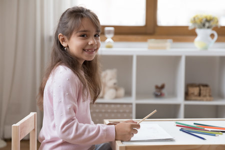 Home portrait of happy adorable little preschool kid girl drawing in paper album, sitting at small desk in playroom, using colorful pencils, looking at camera with toothy gap smileの写真素材