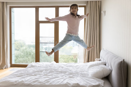 Happy carefree little child jumping on bed mattress at home, flying in air with open arms and legs, laughing, smiling, playing active games, enjoying activities, leisure, motion, having fun aloneの写真素材