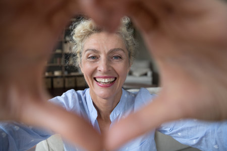 Cheerful pretty mature woman looking at camera through greeting finger heart, making romantic hand...