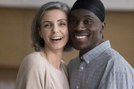 Happy diverse African and Caucasian couple posing indoors, standing close with cheeks touch, looking...