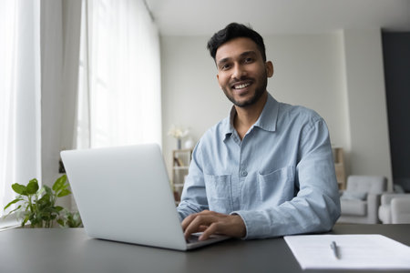 Happy young Indian freelance employee guy working at home, typing on laptop, looking at camera with...