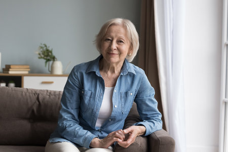 Calm retired mature woman in casual clothes resting alone seated on comfortable sofa in living room smile looking at camera at own modern apartment. Baby-boomer generation female portrait, retirementの写真素材