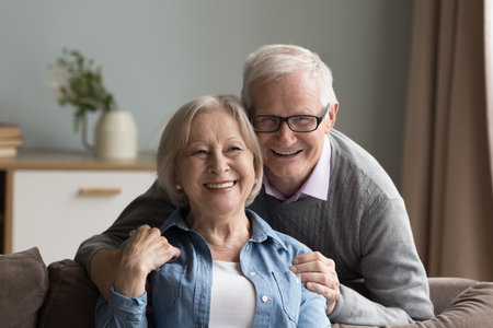 Portrait of happy older 70s grey-haired couple. Mature husband cuddles her senior wife seated on...