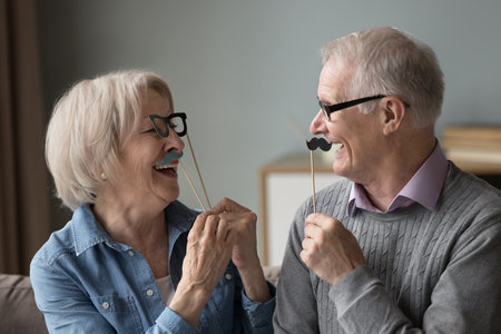 Couple having fun holding party accessories laugh look at each other. Mature family celebrate life event, birthday or anniversary happy strong lifelong marriage. Eyesight check up services for seniorsの写真素材