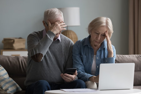 Unhappy old wife and husband sit on couch in living room with laptop and calculator manage family finances, feel desperate due debts, unpaid bills, lack of money to pay monthly bills or bank mortgageの写真素材