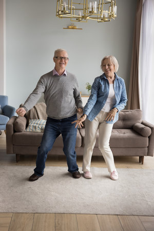Cheerful active old wife and husband have fun in living room together move to rhythm listen favorite music, happy overjoyed senior spouses dance enjoy leisure weekend at new home, feel love to lifeの写真素材