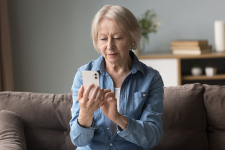 Focused woman read sms on cellphone, share messages use mobile application, learn new app, making call sit on couch in living room. Older generation using modern tech, comfortable remote communicationの写真素材