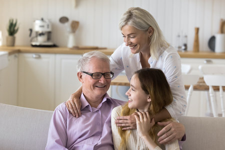 Cheerful grandma and grandpa hugging teenage kid, sitting on home sofa, talking, laughing, smiling, enjoying family leisure , granddaughter visit, weekend, holidayの写真素材